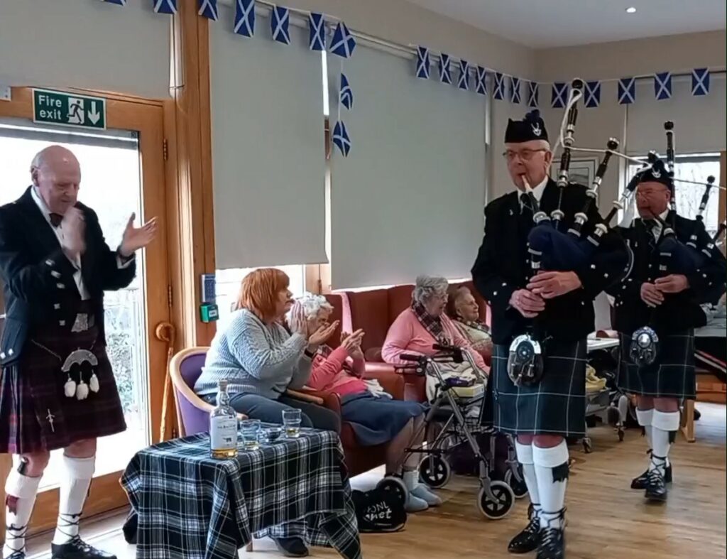 Three men in kilts, one playing the bagpipes, bring Burns Night magic to a room of elderly people beneath Scottish flags. The audience clap along, enjoying a spot of Burns in the festive atmosphere.