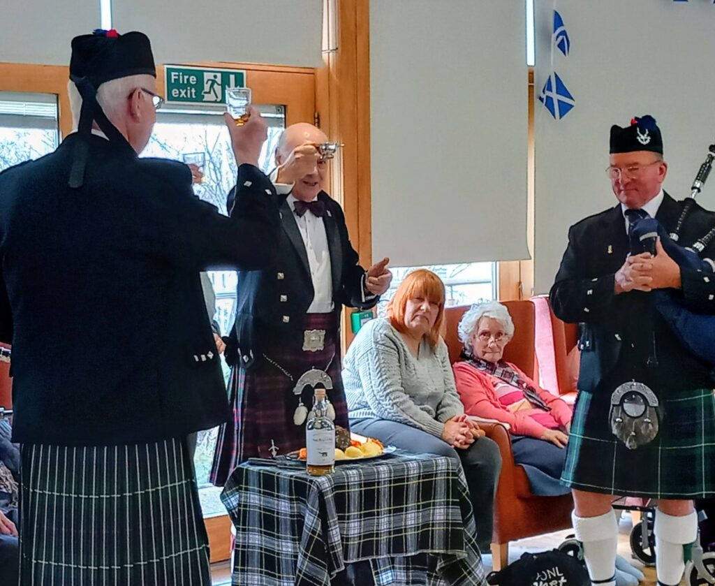 Three men in traditional Scottish dress stand near a table with food and drink, as seated guests observe during a lively Scottish celebration in a room adorned with Scottish flags.