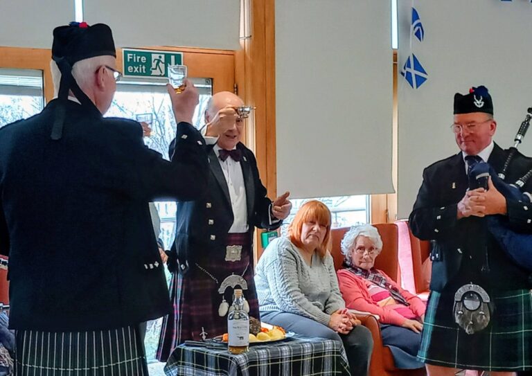 Three men in traditional Scottish dress stand near a table with food and drink, as seated guests observe during a lively Scottish celebration in a room adorned with Scottish flags.