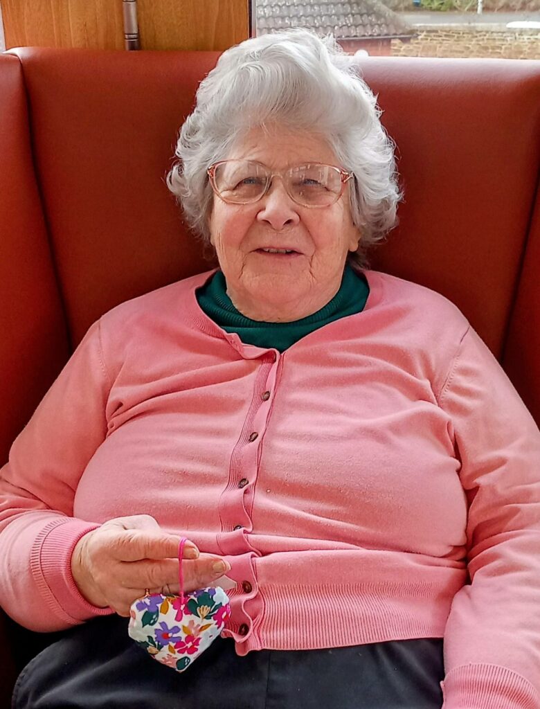 An elderly woman with grey hair, spectacles, and a pink cardigan sits on a red seat, holding a small, floral-patterned heart ornament—her face alight with memories of wagging tails and delightful surprises.