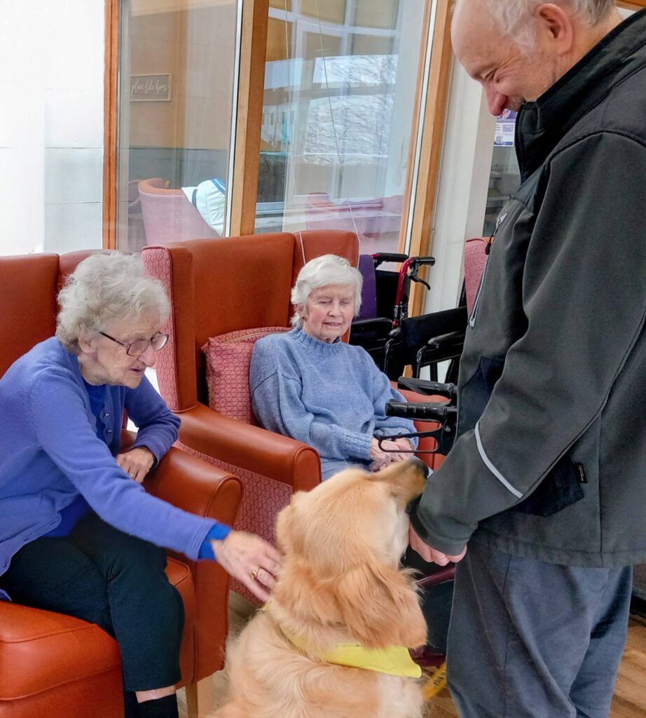 Two elderly women seated in armchairs delight in pleasant surprises as a golden retriever with a wagging tail interacts with them and a standing man in a bright indoor space.
