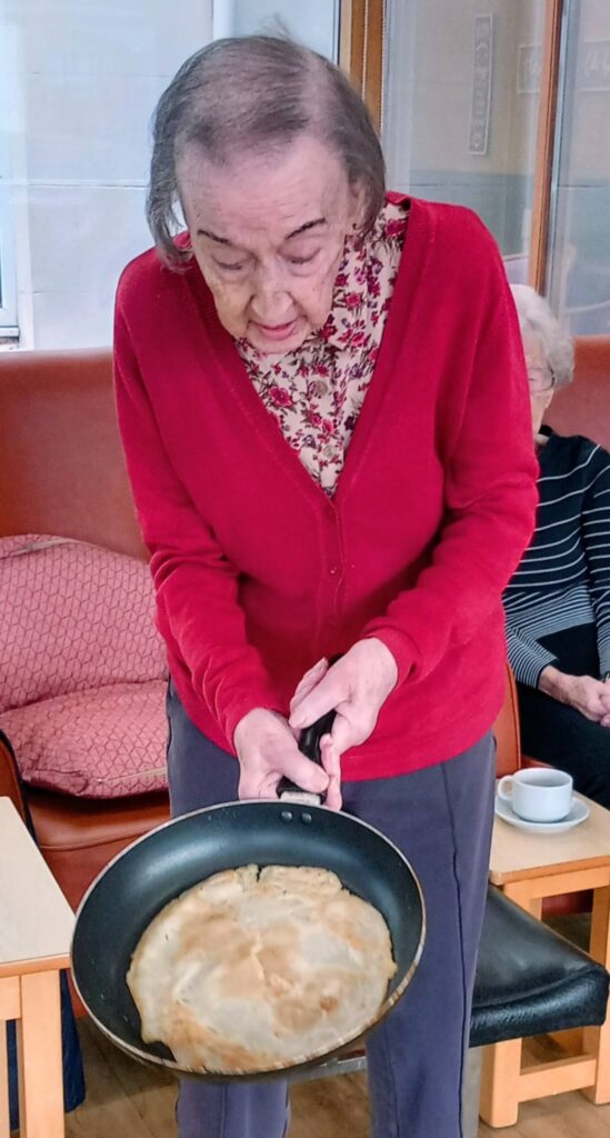 An elderly woman in a red cardigan holds a frying pan with a cooked pancake, poised to flip it, standing indoors near a seated person and beside a table with a cup.