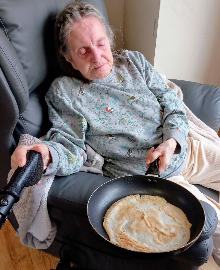 An elderly woman sits in an armchair with a frying pan and a cooked crêpe on her lap, eyes closed and a blanket over her legs—perhaps dreaming of Pancake Day and tossing pancakes.