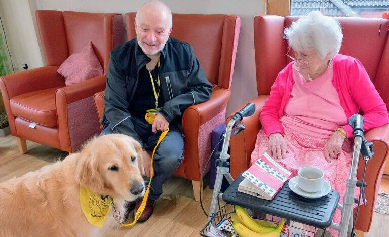 An elderly woman sits in a care home beside a man and a golden retriever in a yellow vest, its wagging tail bringing delightful surprises. A book, cup, and bananas sit on the table in front of her.
