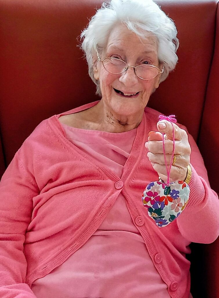 Elderly woman with white hair and glasses, dressed in a pink cardigan, smiles as she holds up a colourful, heart-shaped fabric ornament—her face alight with the delight of lovely surprises.
