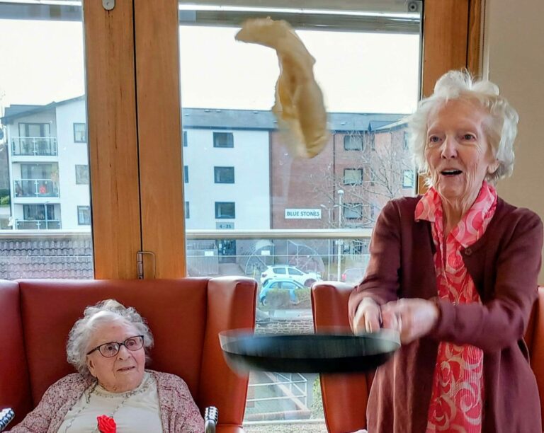 An elderly woman tosses pancakes in a frying pan whilst another elderly woman sits nearby watching in a bright room with large windows, celebrating Shrove Tuesday.