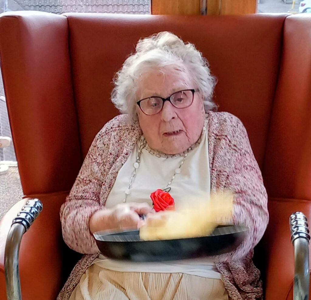 An elderly woman wearing spectacles sits in an armchair, flipping pancakes in a frying pan—possibly trying out her favourite Pancake Day recipes.