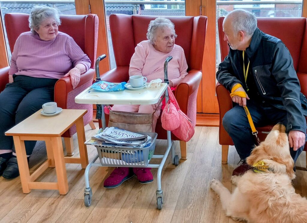 Two elderly women sit in chairs with drinks; one has a walking frame and a tray. Close by, a man with a guide dog, its tail wagging, joins their chat, bringing the prospect of delightful surprises to the afternoon.