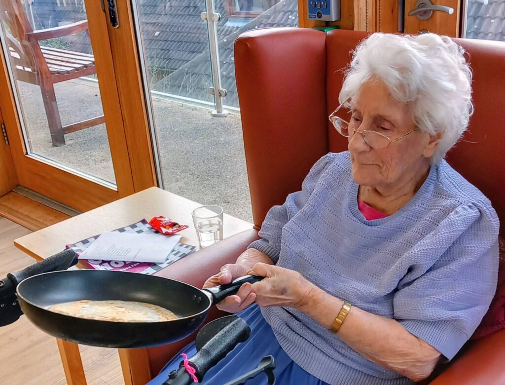 Elderly woman seated in an armchair holding a frying pan with a cooked pancake inside, ready to flip it on Shrove Tuesday, with papers and a glass of water on the table next to her.