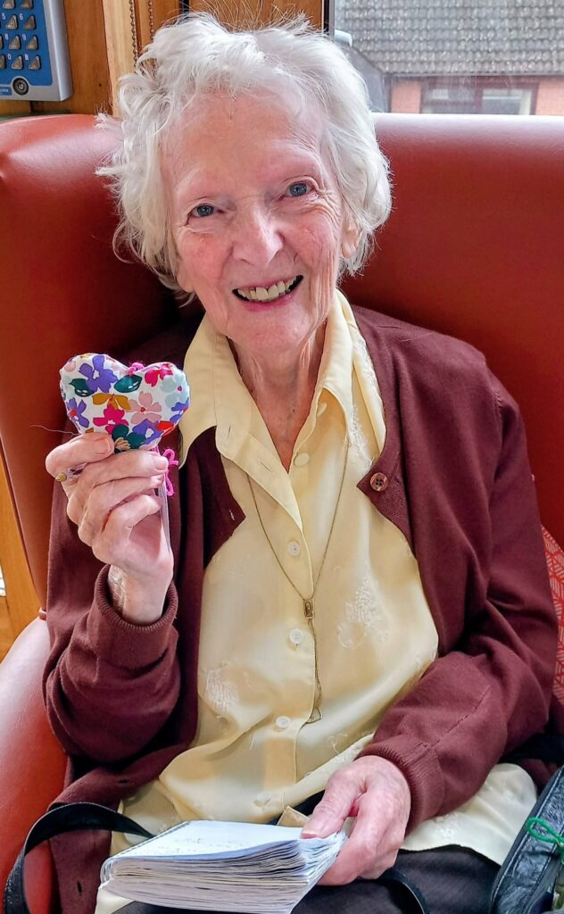 Elderly woman seated in a red chair, smiling and holding a colourful heart-shaped object, with a notebook on her lap—her face radiant with the delight of splendid surprises.