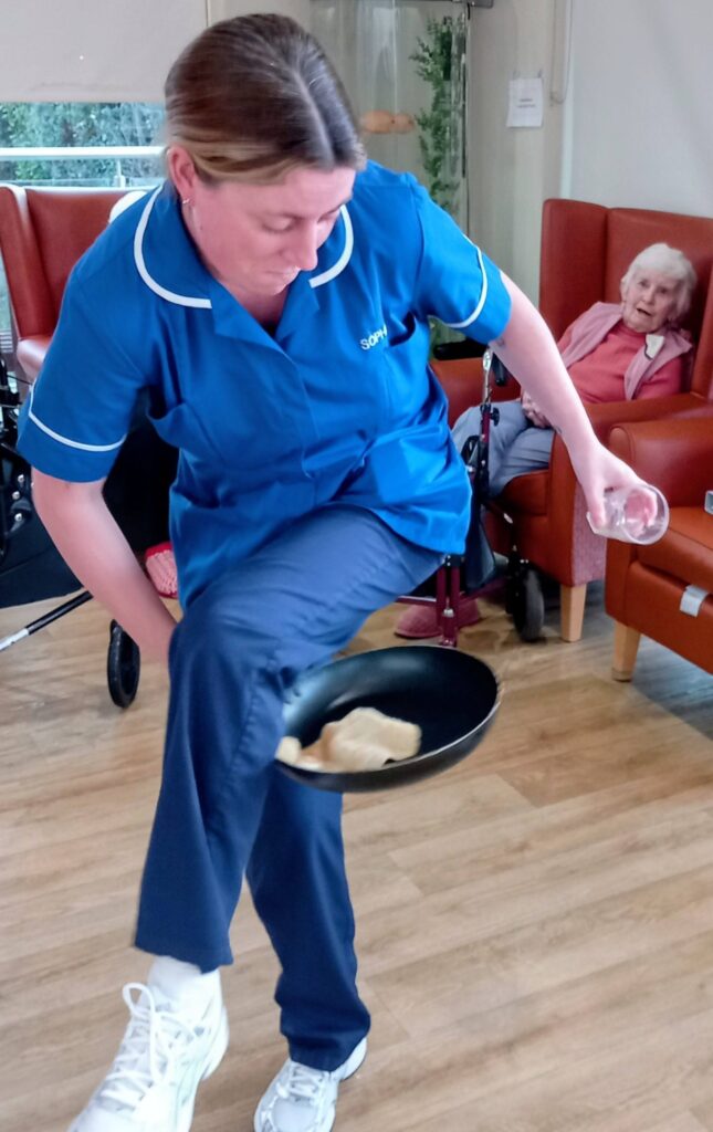 A woman in a blue uniform flips a pancake in a frying pan, demonstrating her pancake tossing skills, whilst an elderly woman looks on from a red armchair in the background.