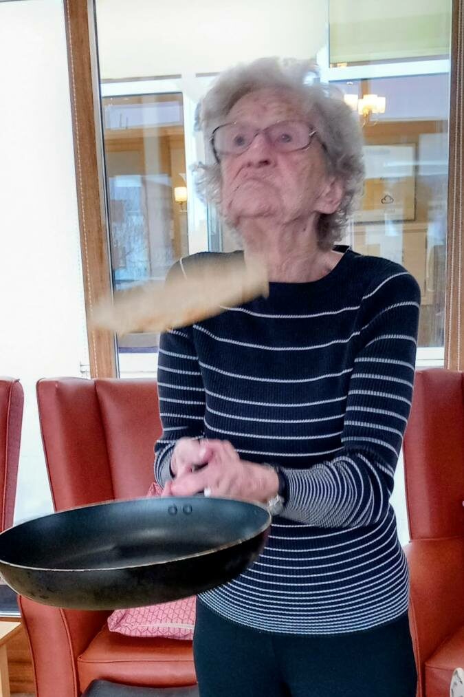 An elderly woman wearing spectacles flips a pancake in a frying pan while standing indoors, celebrating Pancake Day.