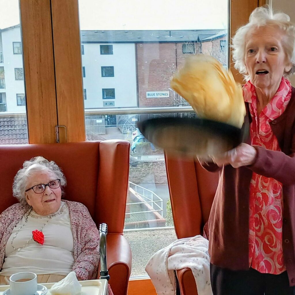 An elderly woman is flipping pancakes in a frying pan while another elderly woman sits nearby, watching. They are indoors with large windows overlooking buildings outside, celebrating Shrove Tuesday together.