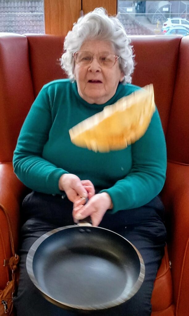 An elderly woman in a green jumper celebrates Pancake Day by tossing a pancake in a frying pan whilst seated on a red chair.