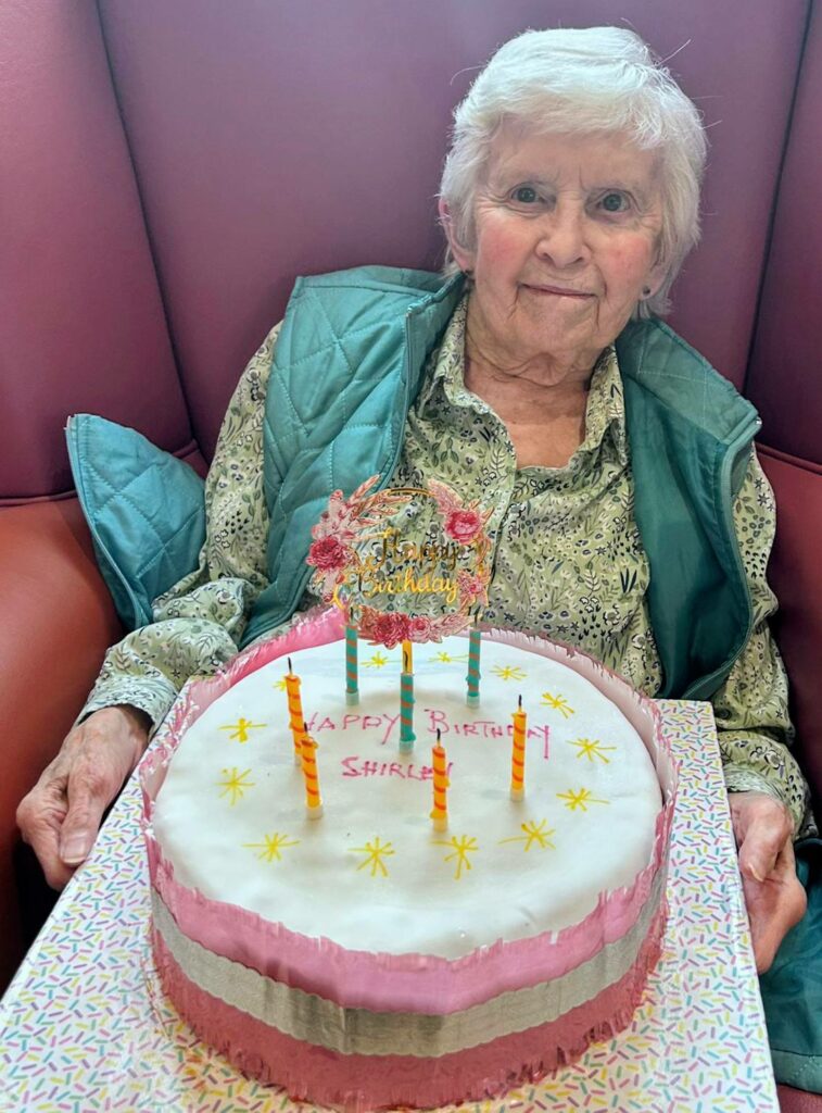 An elderly woman sits in an armchair holding a birthday cake with candles and a message reading Happy Birthday Shirley written on it.