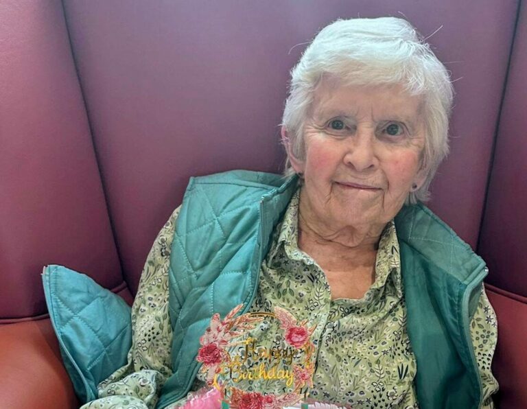 An elderly woman sits in an armchair holding a birthday cake with candles and a message reading Happy Birthday Shirley written on it.
