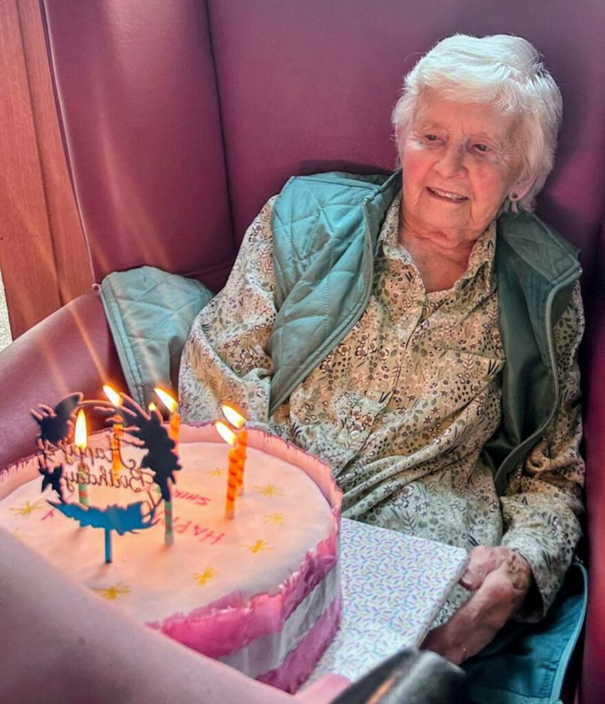 An elderly woman sits in a chair, smiling beside a pink birthday cake with lit candles and a “Happy Birthday” topper.