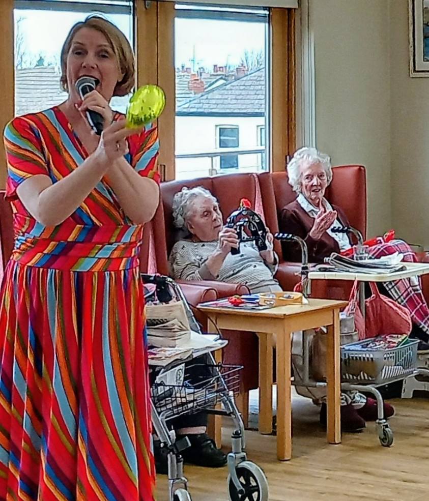 A woman in a colourful striped dress sings into a microphone at a lively Valentine’s Afternoon music event, whilst two elderly women seated behind her watch and hold small musical instruments.