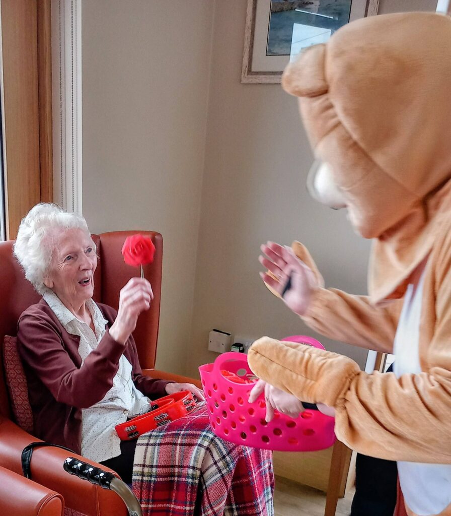 An elderly woman seated in a chair holds a red flower and smiles at someone in a bear costume who is offering a pink basket indoors during a jolly Valentine’s afternoon.