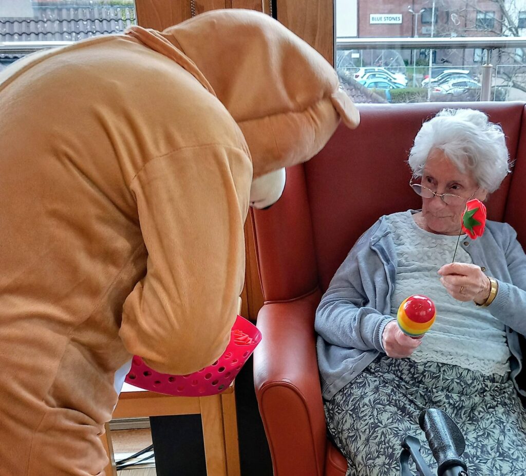 A person in a bear costume presents a pink Valentine’s basket to an elderly woman seated in a chair, who is holding a maraca and a flower-shaped object, bringing a musical touch to the moment.