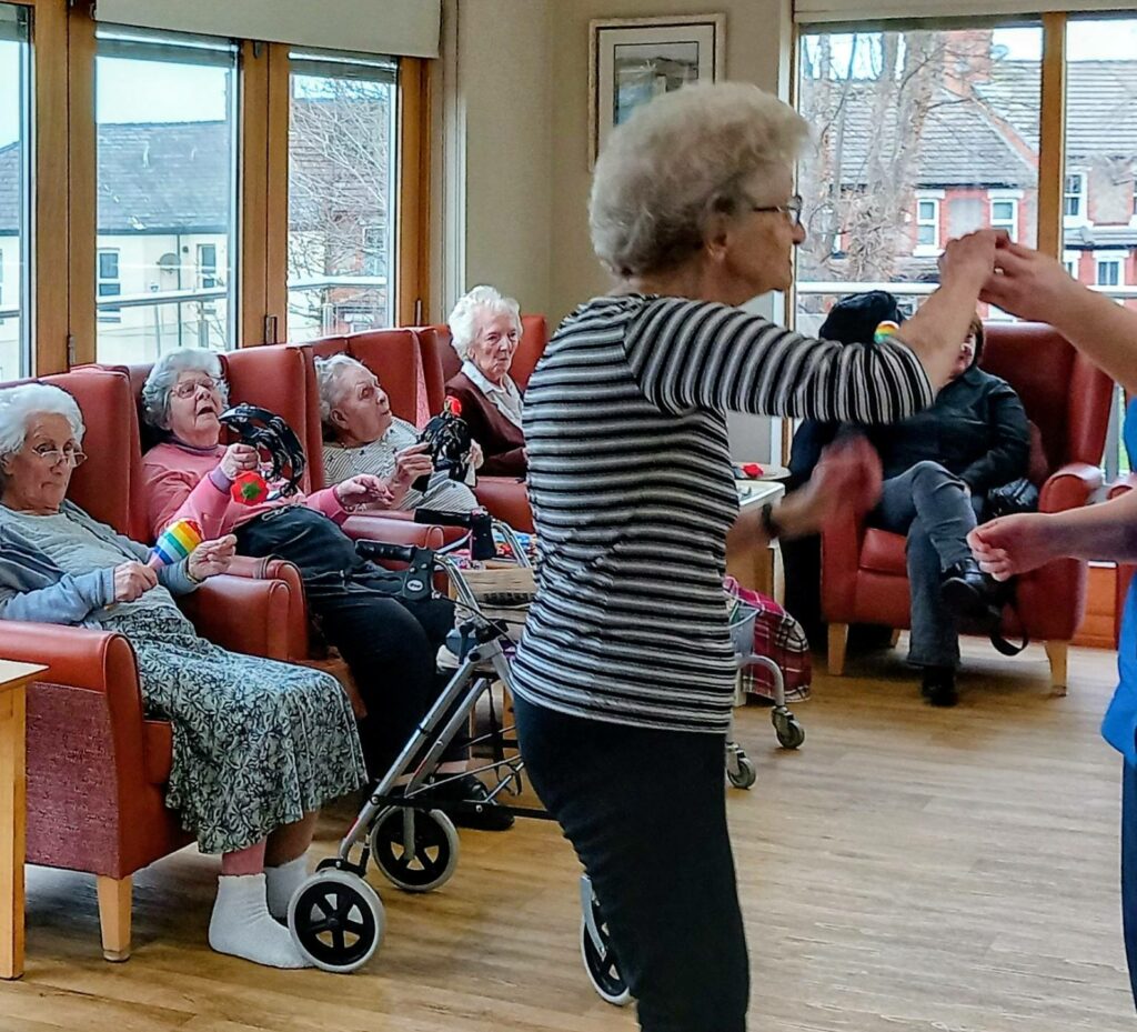 A group of elderly people sit in armchairs in a lounge, enjoying music, whilst one woman stands and dances with another person on a cheerful Valentine’s afternoon.