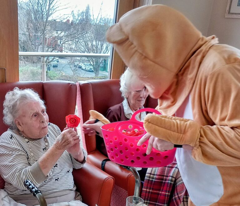 An elderly woman seated in a chair is given a red rose by someone in an animal costume with a pink basket during a Valentine’s Afternoon, as music plays and another elderly woman sits nearby.