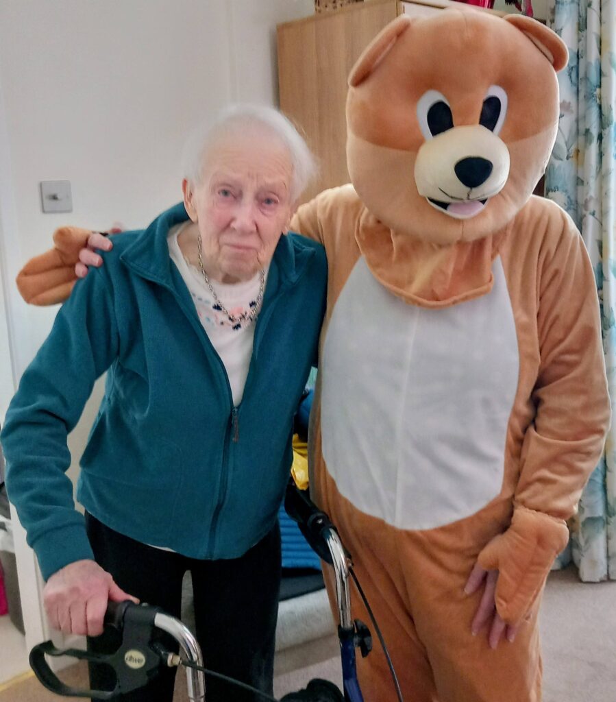 An elderly woman with a walking frame stands beside a person in a bear costume, both facing the camera in a cosy home setting during a jolly Valentine’s afternoon filled with laughter and music.