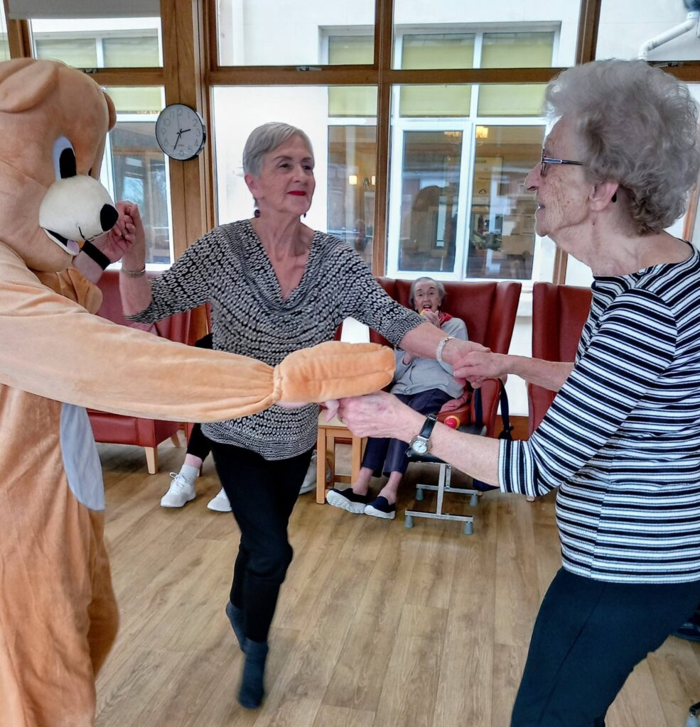 Two elderly women are happily dancing to music with someone in a bear costume in a common room, whilst another lady looks on from a chair in the background during this cheerful Valentine’s afternoon.