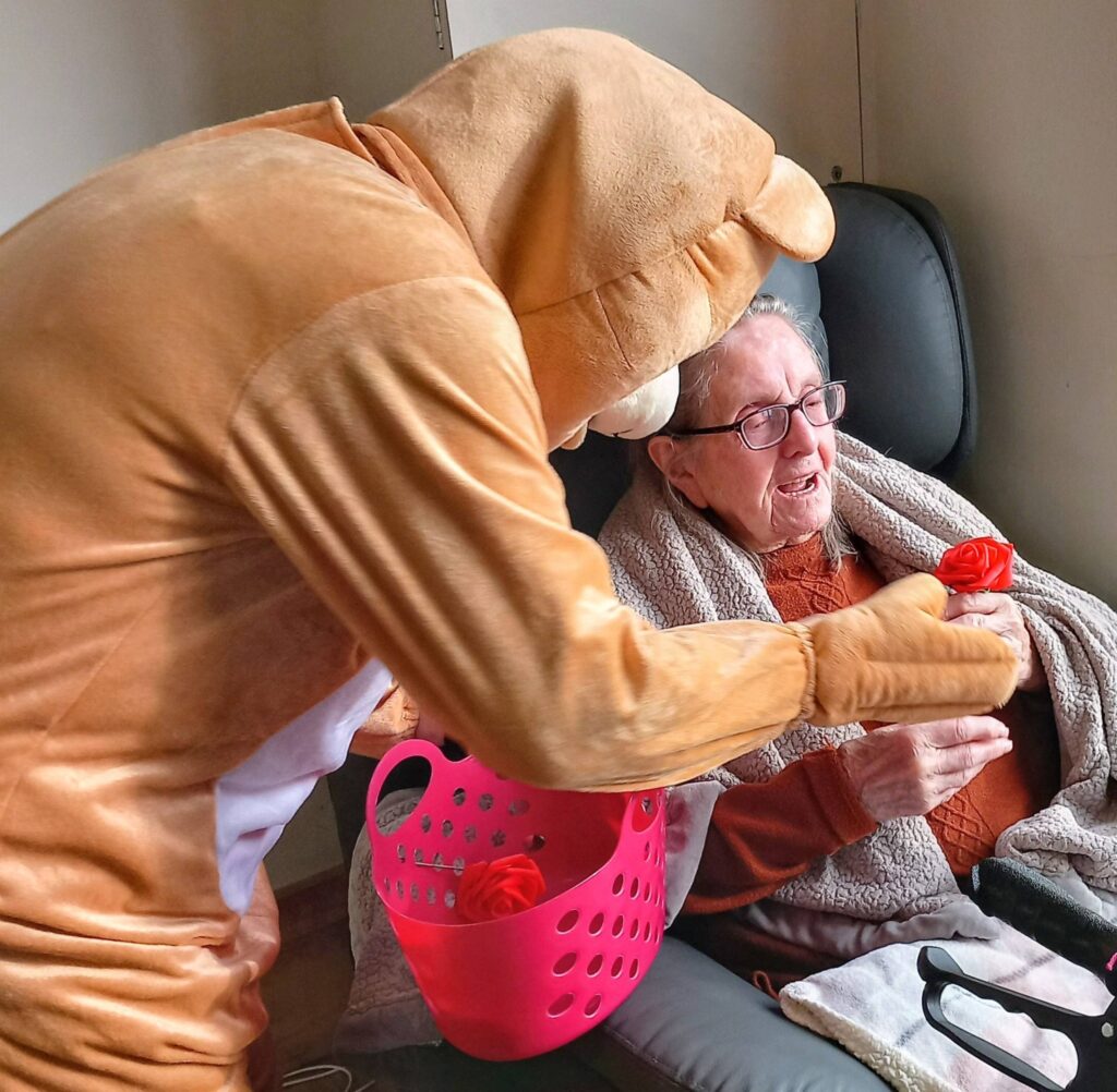A person in a bear costume hands a red rose to an elderly woman seated in a chair, with a pink basket of roses nearby, during a cheerful Valentine’s afternoon filled with dancing and music.