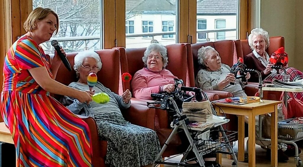 A woman in a colourful striped dress dances with four elderly women seated in armchairs, each holding a soft toy bird, as Valentine’s music fills the bright room with large windows.