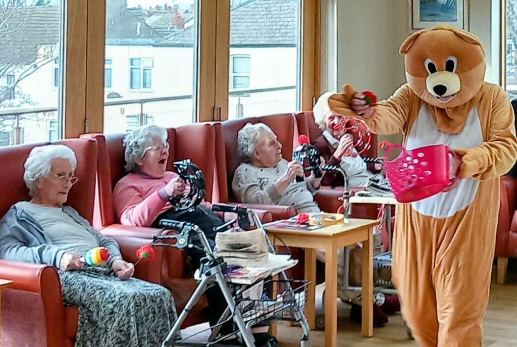A person in a bear costume hands out Valentine’s treats from a pink basket to elderly women seated in a care home lounge, with music gently playing in the background.