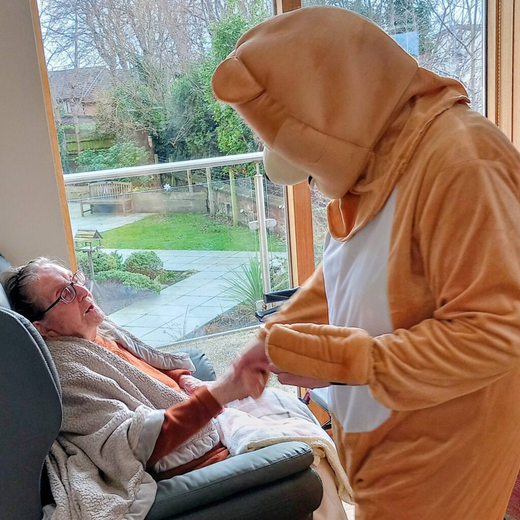A person in a bear costume dances and shakes hands with an elderly person seated in a chair by a window overlooking a garden, bringing music and Valentine’s cheer to this heart-warming scene.