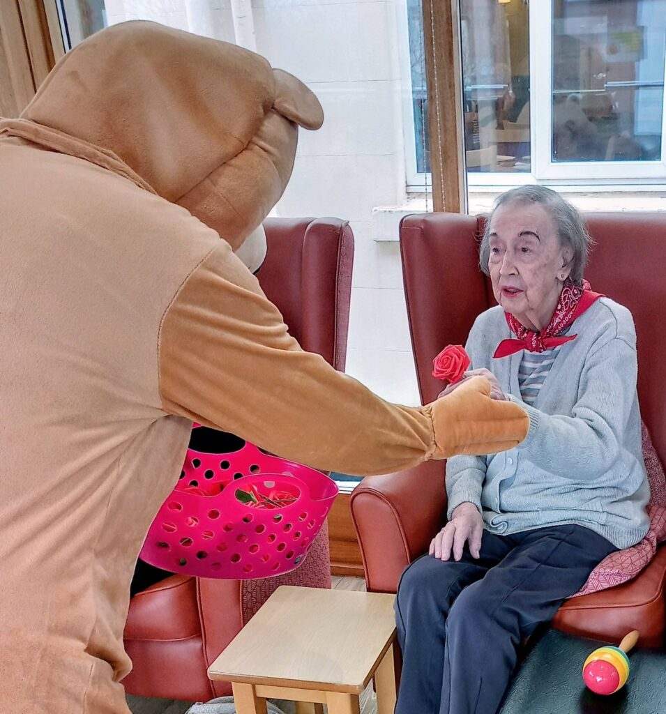 A person in a bear costume hands a red rose to an elderly woman seated in a chair indoors by a window, during a cheerful Valentine’s afternoon filled with music and dancing.