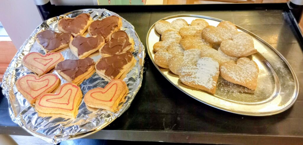 Two trays of heart-shaped biscuits ideal for a Valentine’s afternoon: one tray with chocolate and red icing, the other with plain biscuits lightly dusted with icing sugar.