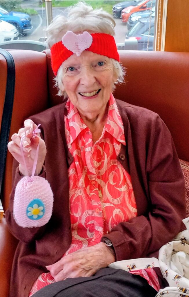 Elderly woman seated on a brown chair, wearing a red hairband with a pink heart, holding a pink knitted pouch adorned with a blue flower, and smiling at the camera.