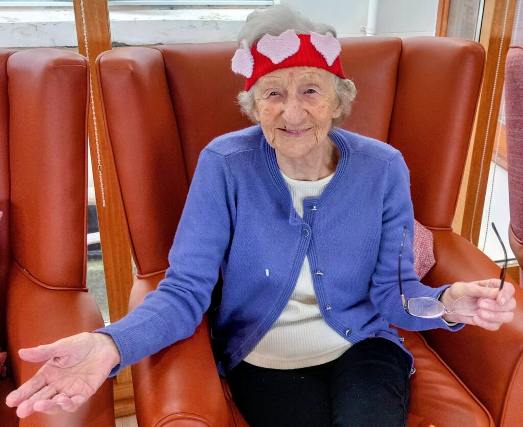 Elderly woman in a blue cardigan sits on a brown armchair, smiling and wearing a red hairband with pink hearts, holding spectacles in her left hand.