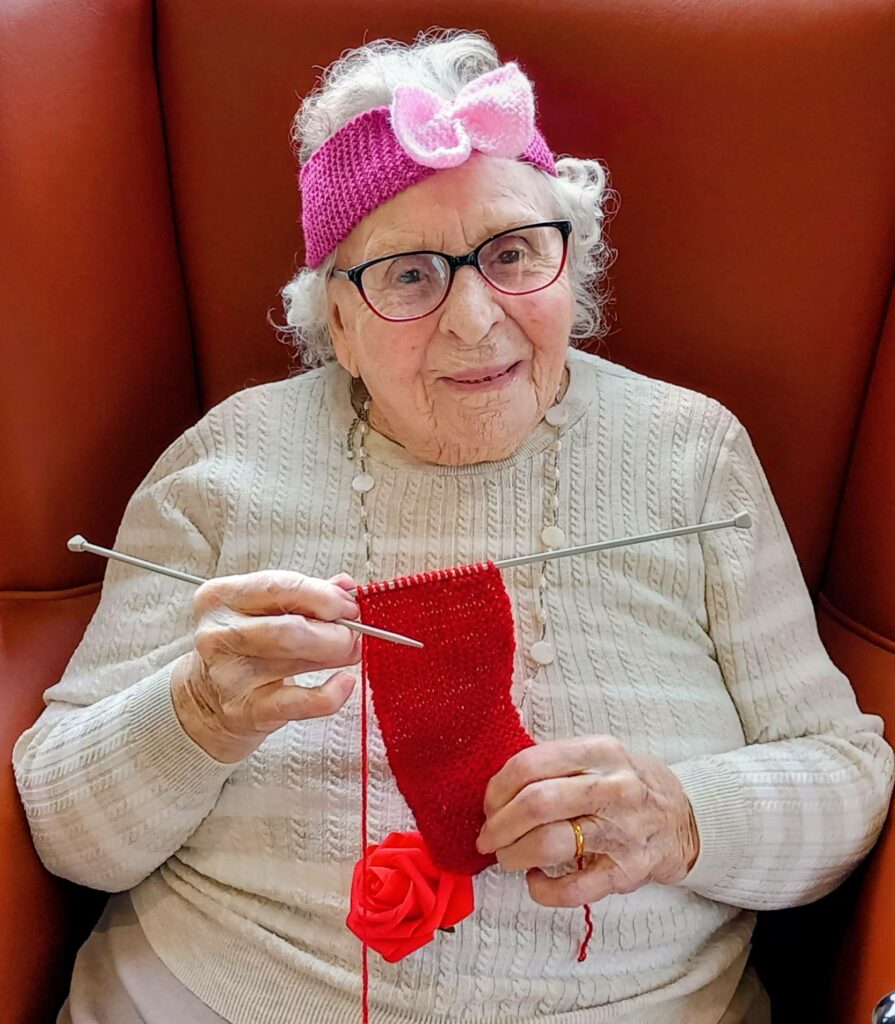 Elderly woman wearing glasses and a pink hairband sits on a red chair, smiling as she knits a red garment with needles.