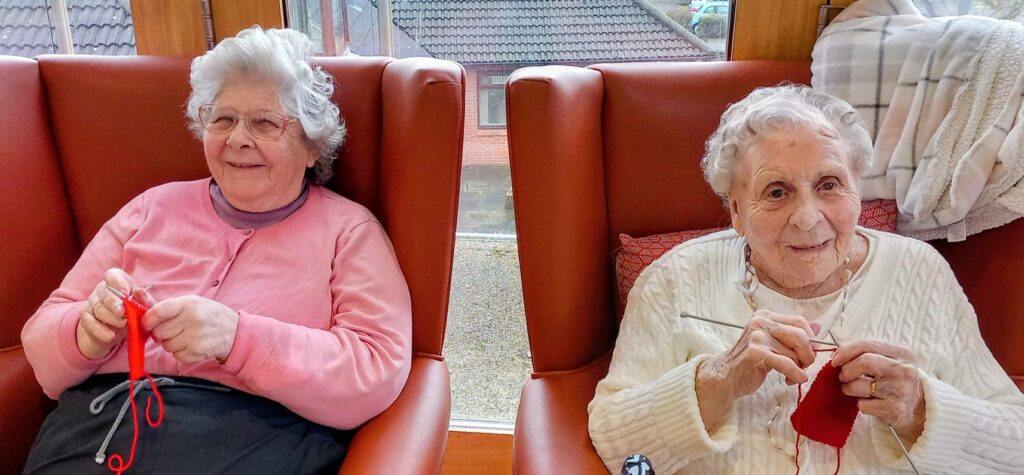 Two elderly women seated in armchairs by a window, both knitting with red wool and smiling at the camera.
