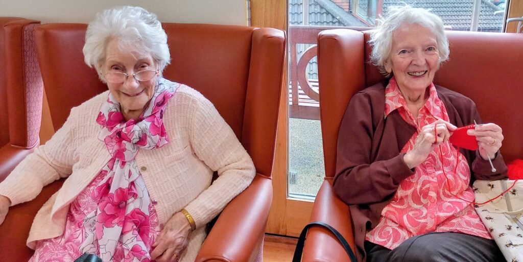 Two elderly women sit side by side in armchairs, smiling; one wears a pink floral scarf, and the other is knitting with red wool.