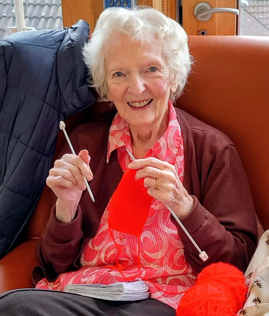 Elderly woman sitting in an armchair, smiling as she knits with red wool and white knitting needles.