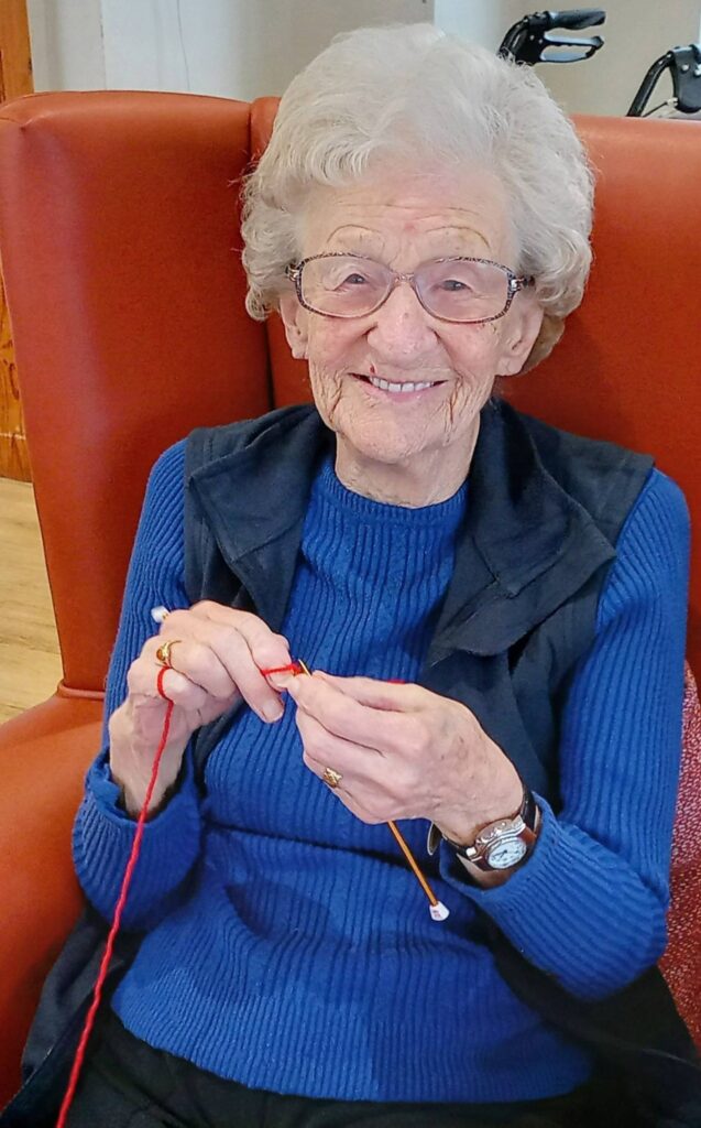 Elderly woman with spectacles sits in an orange armchair, smiling and knitting with red wool.