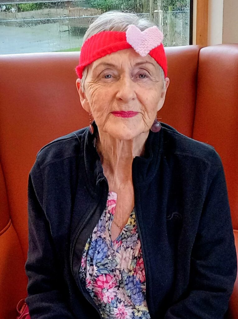 An elderly woman with short grey hair wears a red hairband featuring a pink heart, a black jacket, and a floral top, seated on a brown bench by a window.