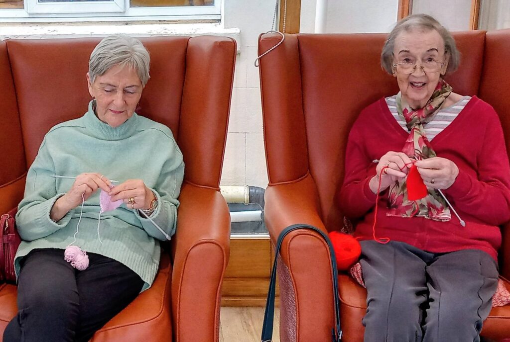 Two elderly women sit in armchairs, knitting with pink and red wool. Both look focused on their craft, with one smiling towards the camera.