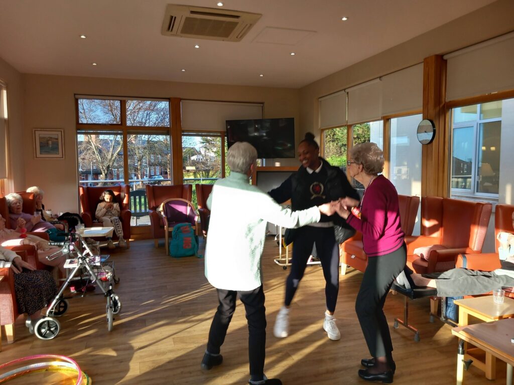 Three women, two older and one younger, are dancing together on a sunny afternoon to "Simply the Best," with people seated and mobility aids visible in the background.