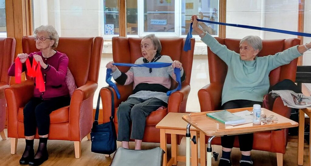 Three elderly women sit in armchairs doing resistance band exercises on a bright afternoon, with a table nearby holding papers and a bottle—simply the best way to remain active together.