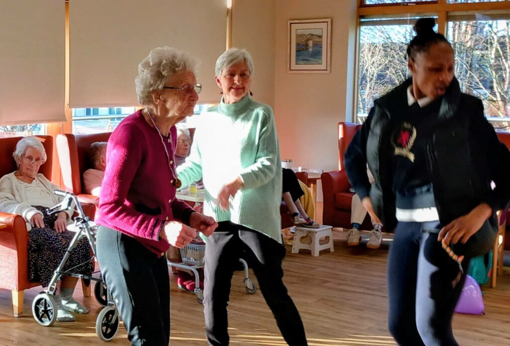 Several women, including two elderly ladies, are dancing to "Simply the Best" in a sunlit room on a cheerful afternoon, with seated pensioners and a walking frame visible in the background.
