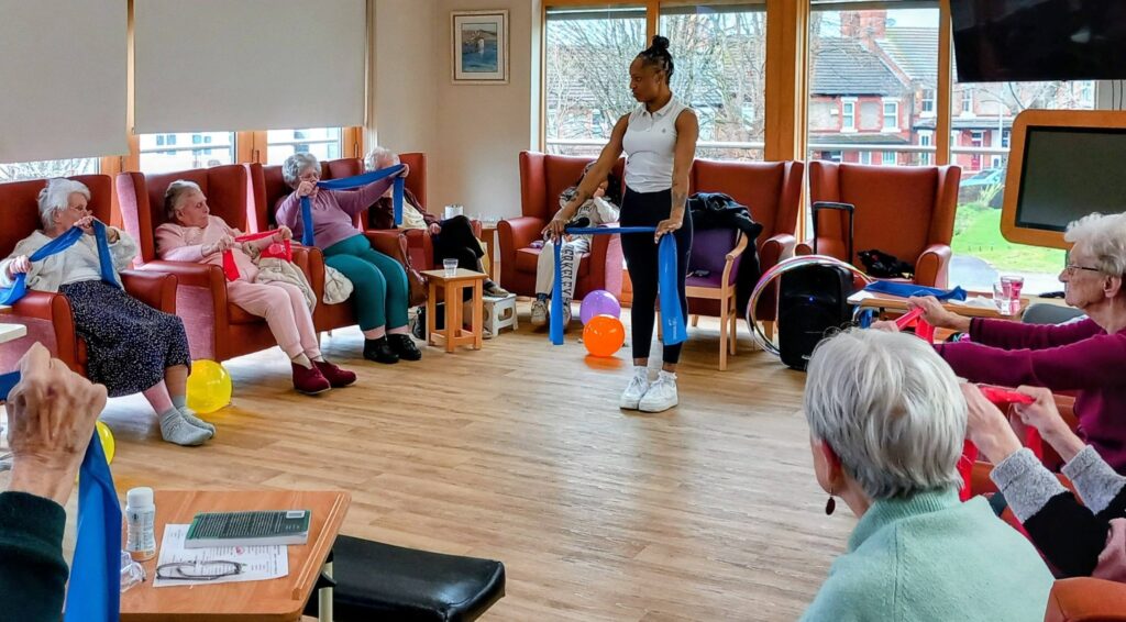 A group of elderly people seated in a circle use resistance bands while an instructor leads a Simply the Best exercise class on a bright afternoon in a room with large windows.