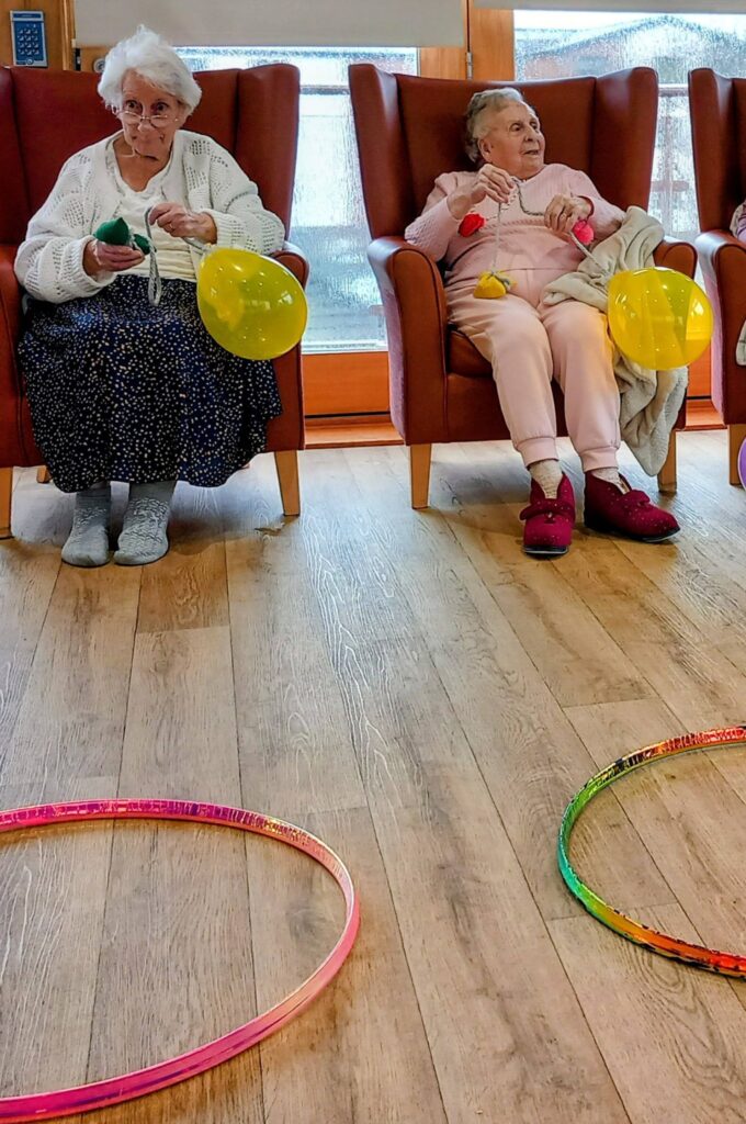 Two elderly women sit in armchairs holding yellow balloons, with colourful hula hoops on the floor in front of them—enjoying simply the best afternoon together.