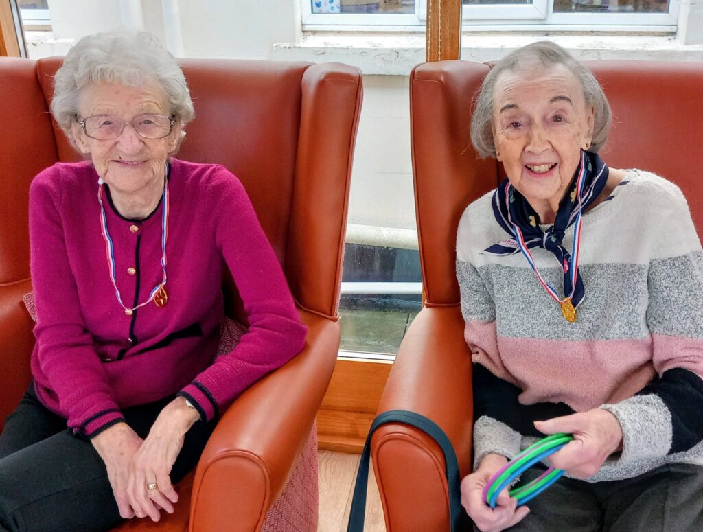 Two elderly women sit in armchairs on a sunny afternoon, smiling at the camera. Both wear medals round their necks—simply the best—while one proudly holds colourful rings in her hands.