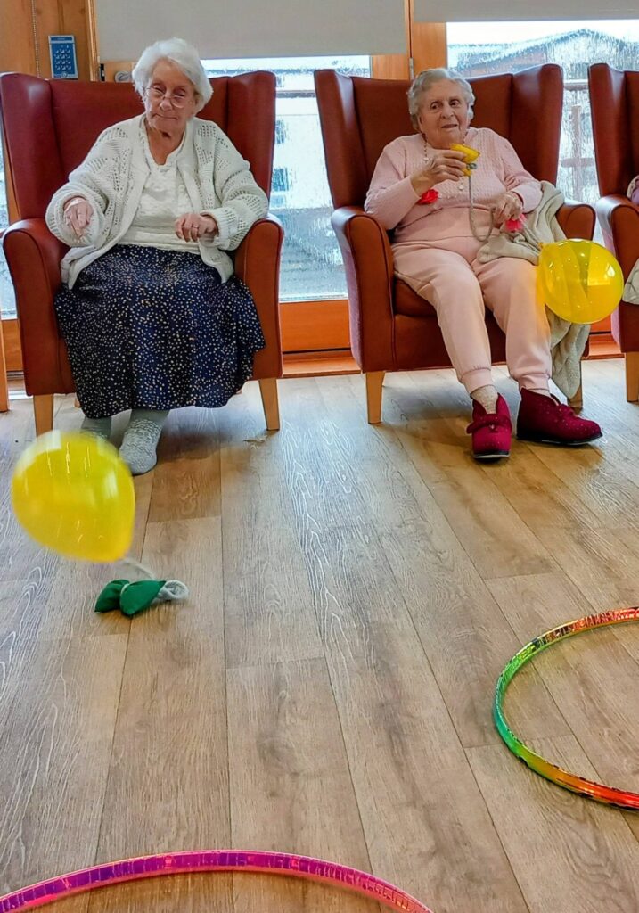 On a cosy afternoon, two elderly women sit in armchairs, each holding a yellow balloon; another balloon and colourful hoops lie on the wooden floor—truly the best way to spend time together.